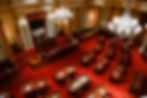 Empty ornate senate chamber with red carpet, desks, chandeliers, U.S. flag, and wall text SENATORIS EST CIVITATIS LIBERTATE TUERI.