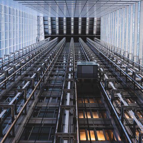 Upward view of a modern glass-and-steel elevator shaft facade with repeating rails and windows, lit in cool blue tones.