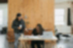 Two people in a cozy workspace. One stands with a coffee, discussing ideas. The other writes at a desk with a laptop, wooden wall behind.