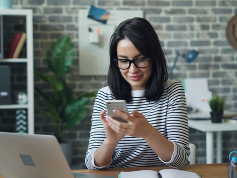 Woman in striped shirt smiles at phone in office with brick wall, laptop, and plants. Cozy and focused atmosphere.