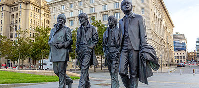 A bronze statue of four men walking side by side is displayed outdoors in an urban setting, with historic buildings and trees in the background.