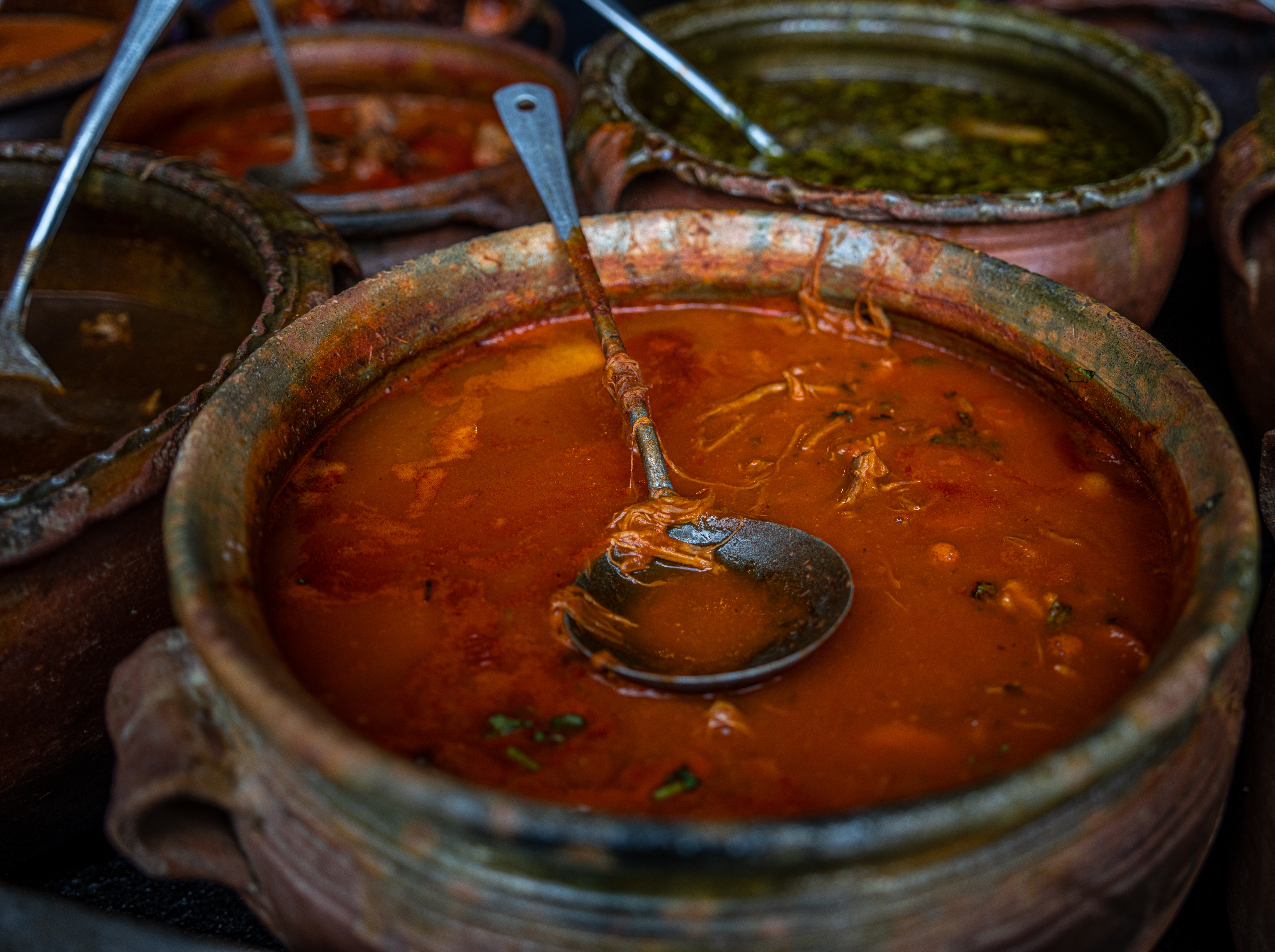 Clay pot filled with Moroccan harira soup, featuring a metal ladle. Surrounding pots contain different colored stews. Earthy setting, rustic vibe.