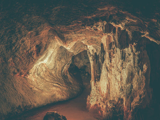 Caverna iluminada com paredes rochosas marrons e laranjas. O chão é de terra escura. A atmosfera é misteriosa e tranquila.