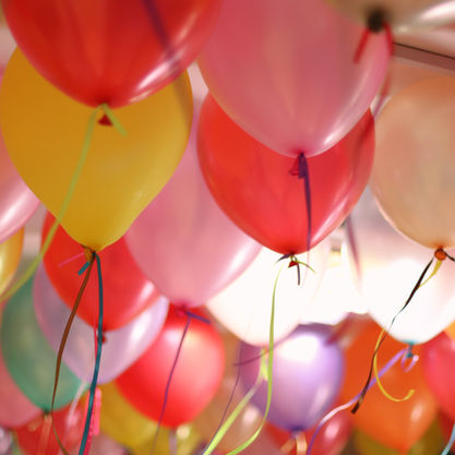 Colorful balloons with ribbons fill the ceiling, creating a festive and joyful ambiance. Bright reds, yellows, and pinks dominate the scene.