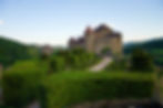 Medieval castle with turrets, surrounded by manicured topiary gardens. Rolling green hills in the background under a clear blue sky.