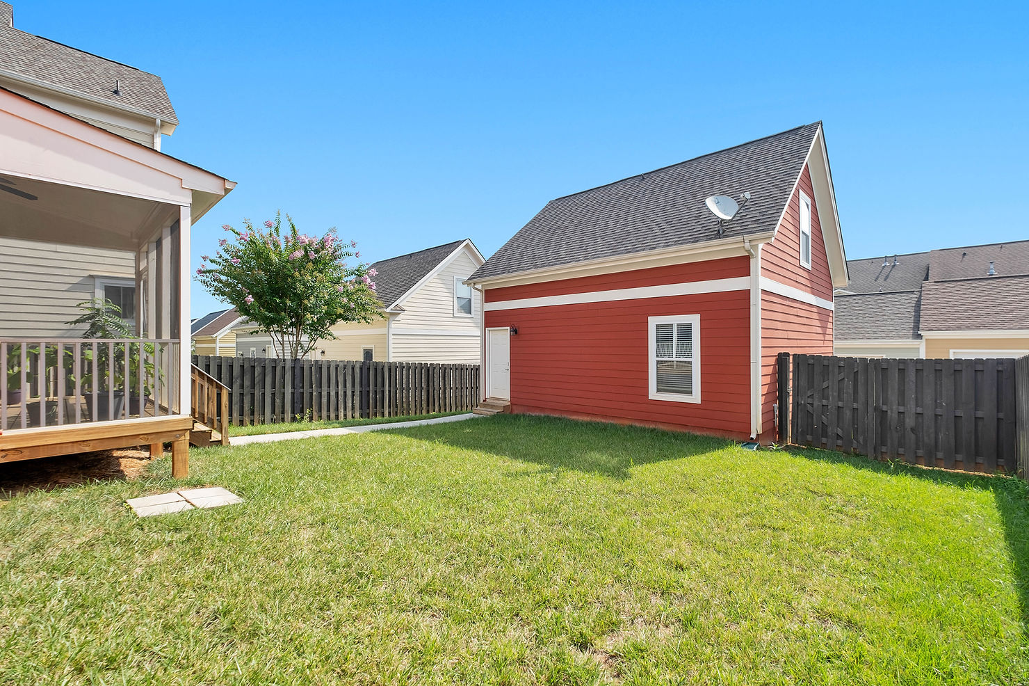 A well-maintained backyard enclosed by a tall, dark wooden fence, with a red detached garage, a light-coloured house with a screened porch, a small paved path and a grassy area under a clear blue sky.