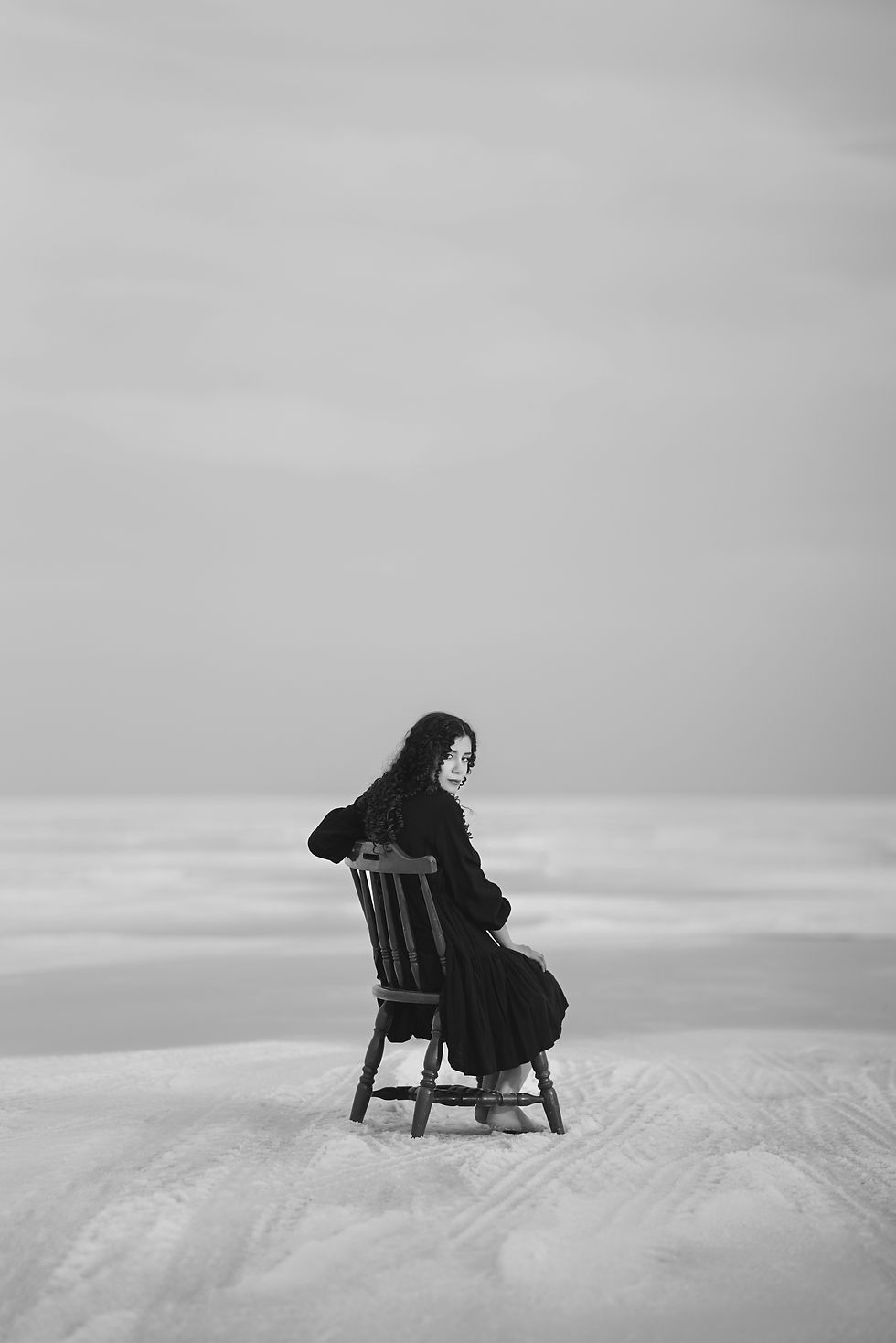 Person in black dress sits on a chair in a snowy landscape, looking back. The scene is grayscale, with a serene and contemplative mood.