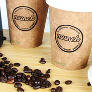 Two brown "munch" cups on a wooden surface, surrounded by coffee beans, a black coffee maker, and chocolate chip cookies. Cozy cafe vibe.