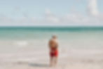Man in red shorts stands on a beach, facing the turquoise sea under a partly cloudy sky, hands behind his back, creating a serene mood.