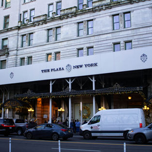 The Plaza New York hotel facade with parked cars and a white van under festive lights at the entrance, city street scene