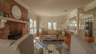 Cozy open-plan living room with brick fireplace, beige sofas, glass coffee table, patterned rug, and kitchen bar in back.