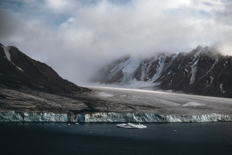 Snow-covered mountains with mist, a glacier descending into the ocean, and an iceberg in the foreground. Moody, cloudy sky overhead.