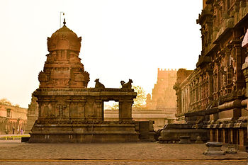 A carved stone shrine pavilion stands in the foreground of a vast temple courtyard in Tamil Nadu, India, bathed in warm golden light at dusk, with a towering gopuram gateway visible in the background against a hazy amber sky.