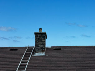 Rooftop with a chimney and ladder against a clear blue sky. The roof is covered with dark shingles. A few small clouds float in the background.
