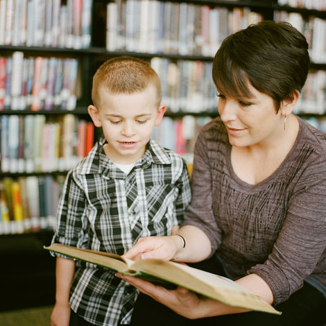 Teacher and young learner in library reading and looking at a book.