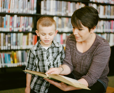 Teacher and young learner in library reading and looking at a book.