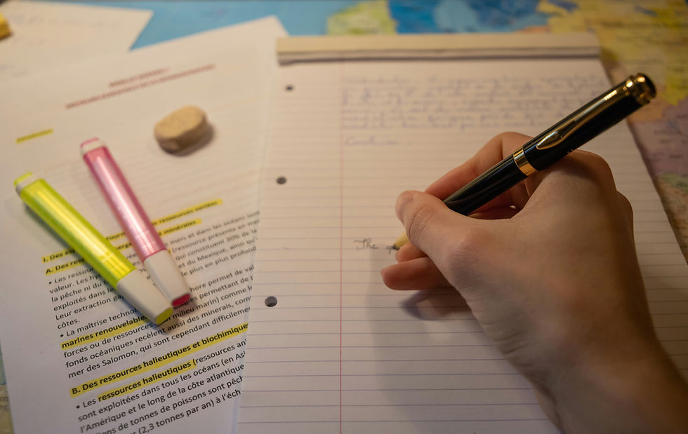 Hand writing on lined paper with black pen. Highlighters and typed notes are visible. Background shows a map, creating a studious mood.