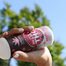 Person drinking from a red and white Delta 9 THC grapefruit drink can outdoors with a clear blue sky and green trees in the background.