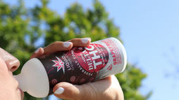 Person drinking from a red and white Delta 9 THC grapefruit drink can outdoors with a clear blue sky and green trees in the background.