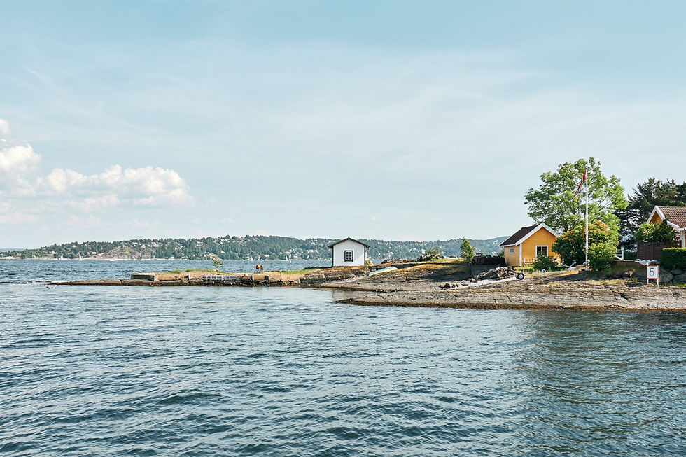 Calm seaside with small houses, one yellow, on rocky shore. Clear blue sky and water, lush greenery. A peaceful, idyllic scene.