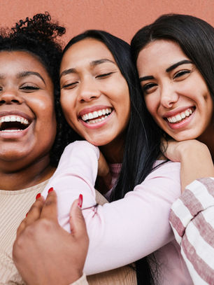 Three smiling women hug close together against a peach wall, wearing beige, pink, and striped tops, joyful and relaxed.
