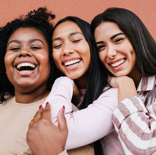 Three women smiling joyfully, embracing each other. Background features a warm, terracotta wall. One wears stripes, others in pastels. Warm mood.