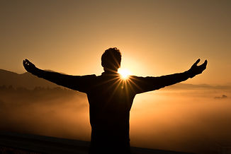 man facing sunrise on top of a mountain above the clouds with the end of twighlight leaving the sky and new day begins. Nao Wellbeing supports individuals seeking change, this is for you. Man's arms open embracing a new day.