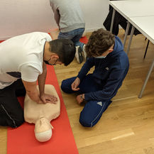 Two people practice CPR on a mannequin in a classroom setting during BLS training. They are focused and wearing masks, with red mats on a wooden floor.