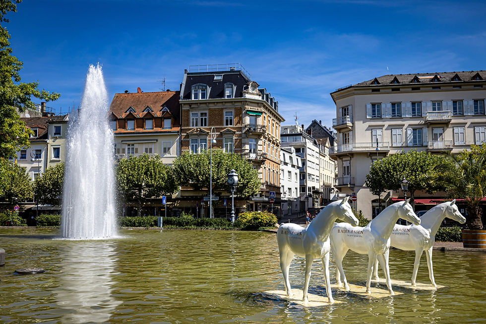 Fontaine jaillissante avec sculptures de chevaux blancs sur l'eau, entourée de bâtiments anciens et arbres verdoyants sous ciel bleu.