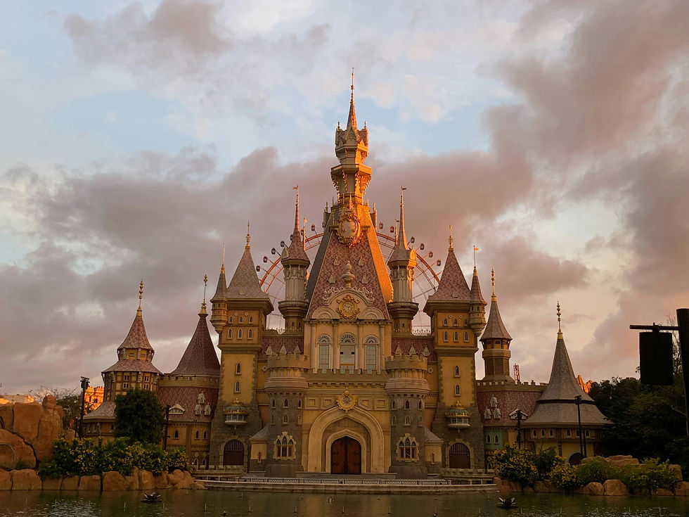 Fairytale castle at sunset, with pink towers and gothic spires, surrounded by water and greenery, under a cloudy sky.