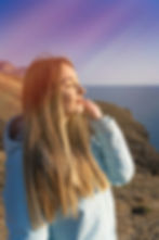 Woman in a light blue jacket with long hair enjoys the sun on a cliff by the sea, eyes closed, serene expression, clear sky background.
