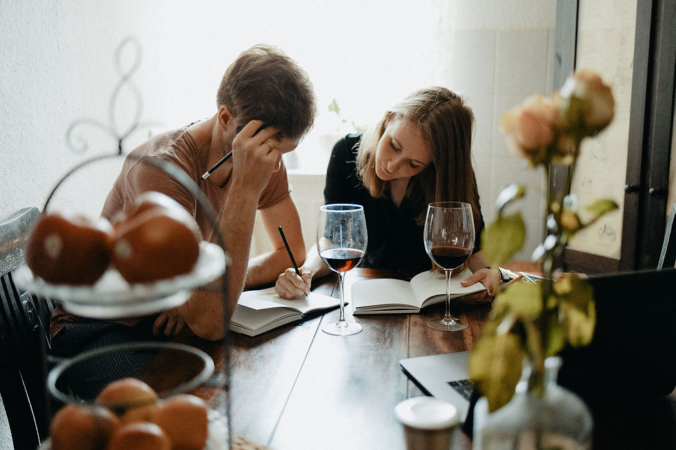 Two people sit at a wooden table, writing and studying. There are open books, wine glasses, apples, and flowers. Bright and focused mood.