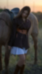 Young woman in a dark jacket leans against a saddled horse in a sunset field, looking calm.