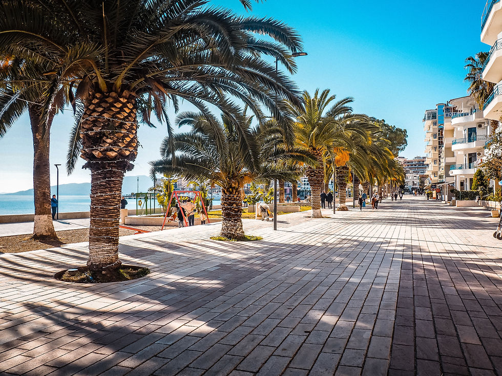 A picture of the Saranda promenade, featuring a wide sidewalk, palm trees, and the sea on one side. People are walking, and some bikes are parked along the way.