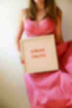 Woman in a pink dress holds a sign reading "SPEAK TRUTH" against a plain background. The mood is empowering.