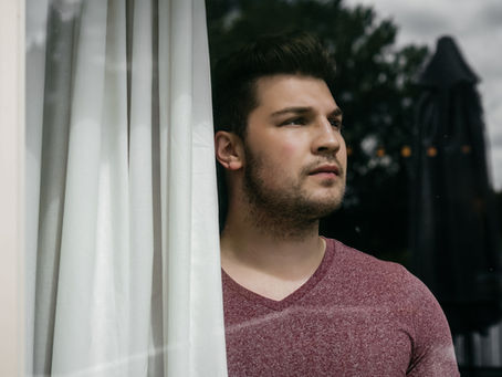 Thoughtful man in a maroon shirt looks out a window beside white curtains, with blurred trees and patio behind him.