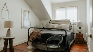Cozy bedroom with a black metal bed, gray bedding, wooden floor, and simple decor. Two lamps and a potted plant beside the bed. Bright window.