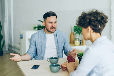 Two people sitting at a table in a kitchen, talking intently. Two teal mugs, a smartphone, and red apples are on the table. Plants in background.