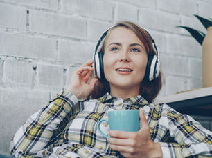 Smiling woman in headphones relaxes on a couch, holding a blue mug, against a white brick wall.