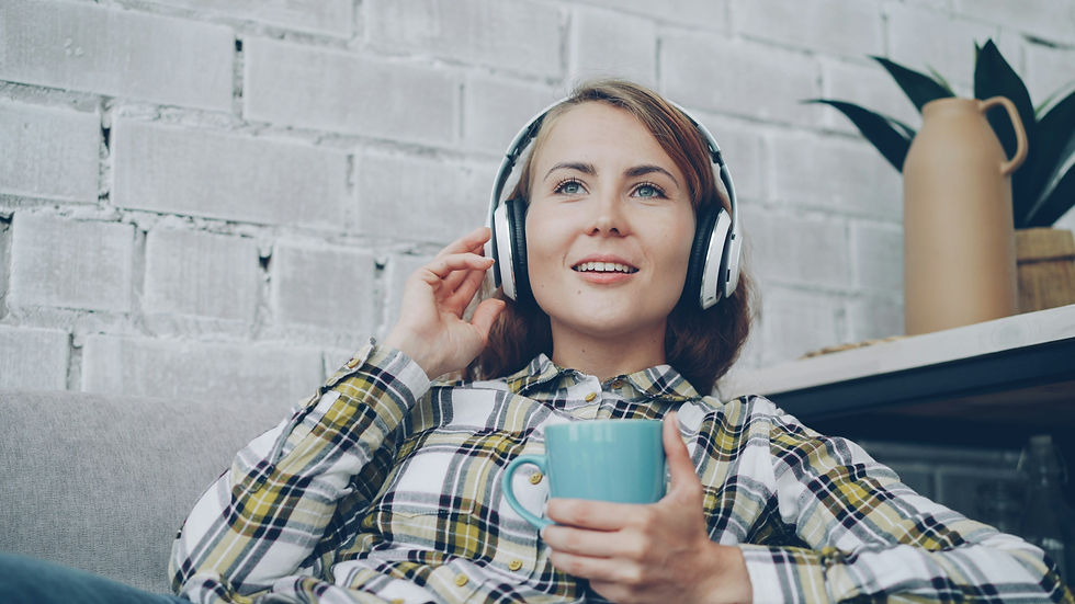 Smiling woman in headphones relaxes on a couch, holding a blue mug, against a white brick wall.