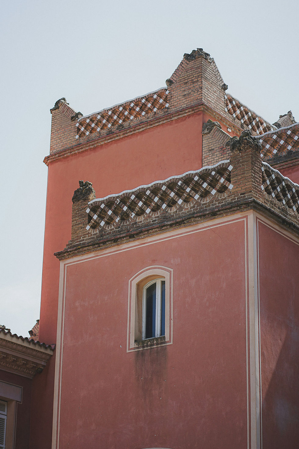 Brick building with pink walls and ornate lattice brickwork at the top. A single arched window is visible. Clear sky background.
