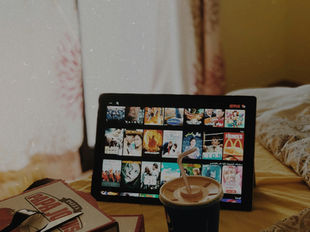 Feet in socks on a coffee table facing a TV displaying "Netflix." Room includes a guitar, plants, books, and a relaxed atmosphere.