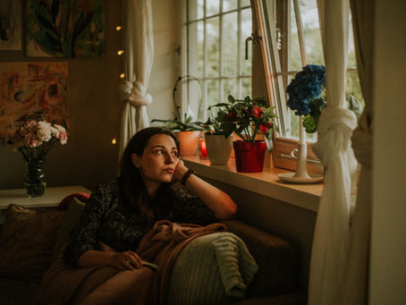 Woman sits by a sunlit window in a cozy room with flowers and candles, gazing pensively outside.