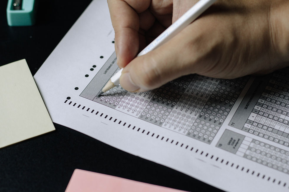 Hand filling a multiple-choice answer sheet with a pencil. The sheet is on a black surface with colored papers visible.