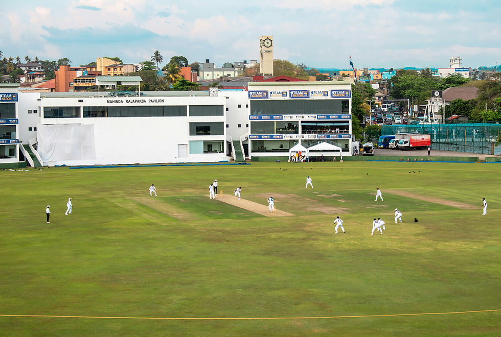 Photo Credit: Zoshua Colah

Cricket match in action on a green field, in front of a white building labeled Mahinda Rajapaksa Pavilion. Urban backdrop with colorful buildings.