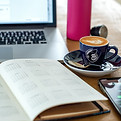 A notebook and a cup of coffee sit on a wooden table in front of a silver laptop.