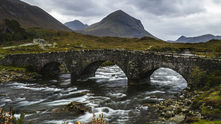 Pont de pierre à trois arches sur une rivière vive, montagnes sombres et ciel d’orage, fleurs violettes au premier plan.