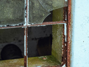 Rusty window frame with cracked glass on an old wall. Visible green moss and chipped paint create a worn, neglected atmosphere.