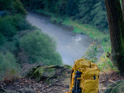 A mustard-yellow hiking backpack rests on a rocky forest overlook above a winding river, symbolizing setting down unnecessary burdens and moving forward with greater perspective.
