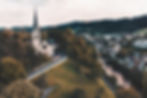 A church with a tall steeple sits on a hill surrounded by trees. A river runs nearby with a town and mountains in the background. Quiet, serene.